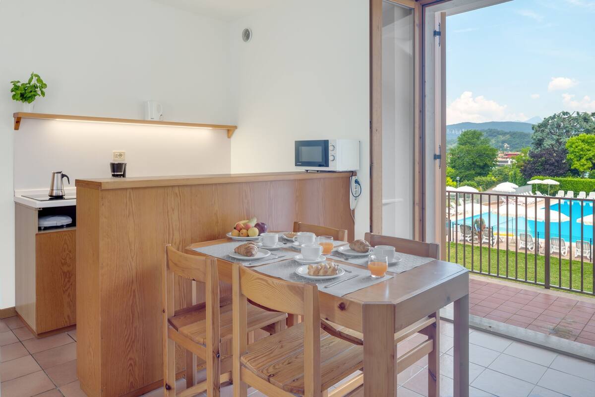 A breakfast setup with a wooden table and chairs, a coffee maker, a microwave, and a view of a swimming pool and greenery.