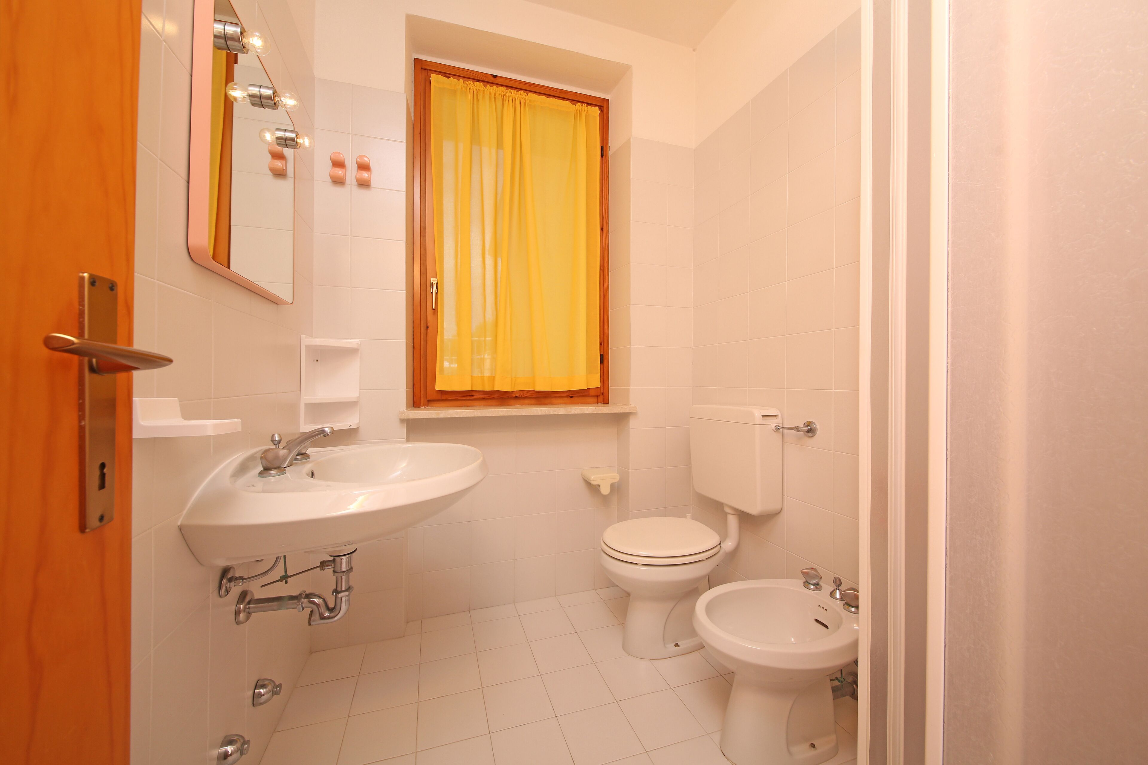 Een badkamer met een wastafel, toilet, bidet en een raam met een geel gordijn.