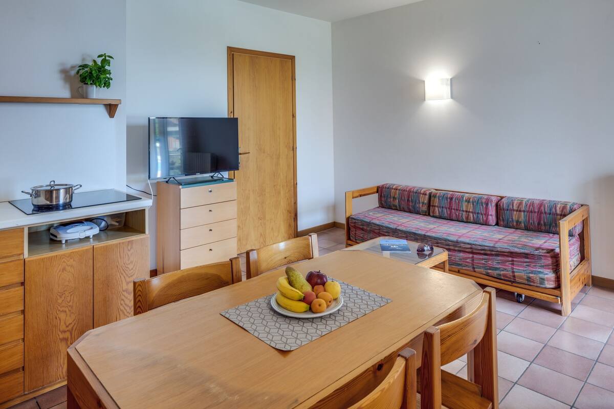 A compact living space with a dining table set with fruit, a sofa, a TV, and a kitchenette.