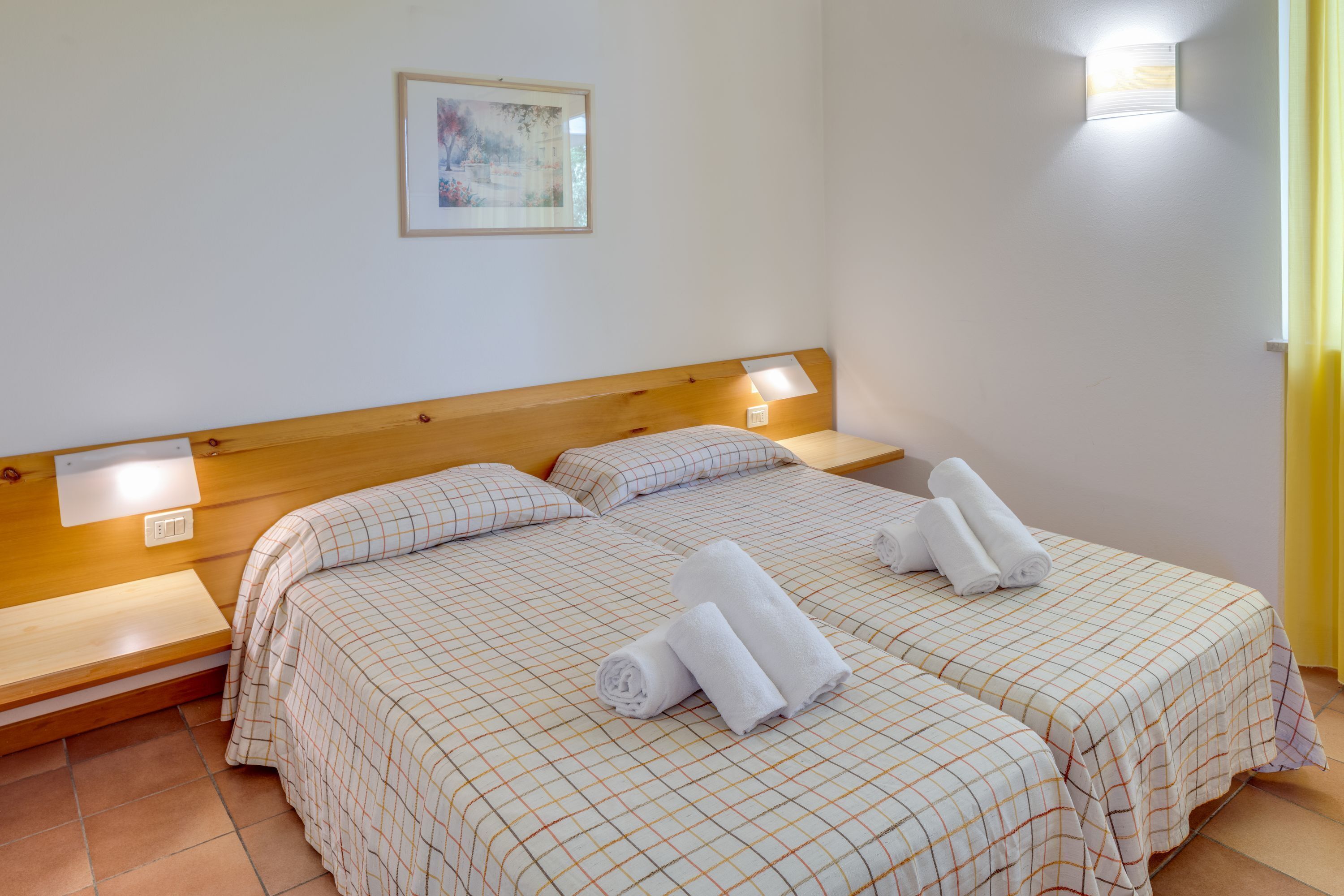 A hotel room with two beds, each with a wooden headboard and a checkered bedspread. There are two bedside tables, a wall-mounted light, and a framed picture on the wall.