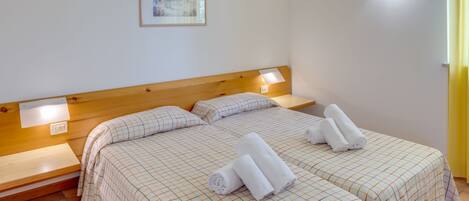 A hotel room with two beds, each with a wooden headboard and a checkered bedspread. There are two bedside tables, a wall-mounted light, and a framed picture on the wall.