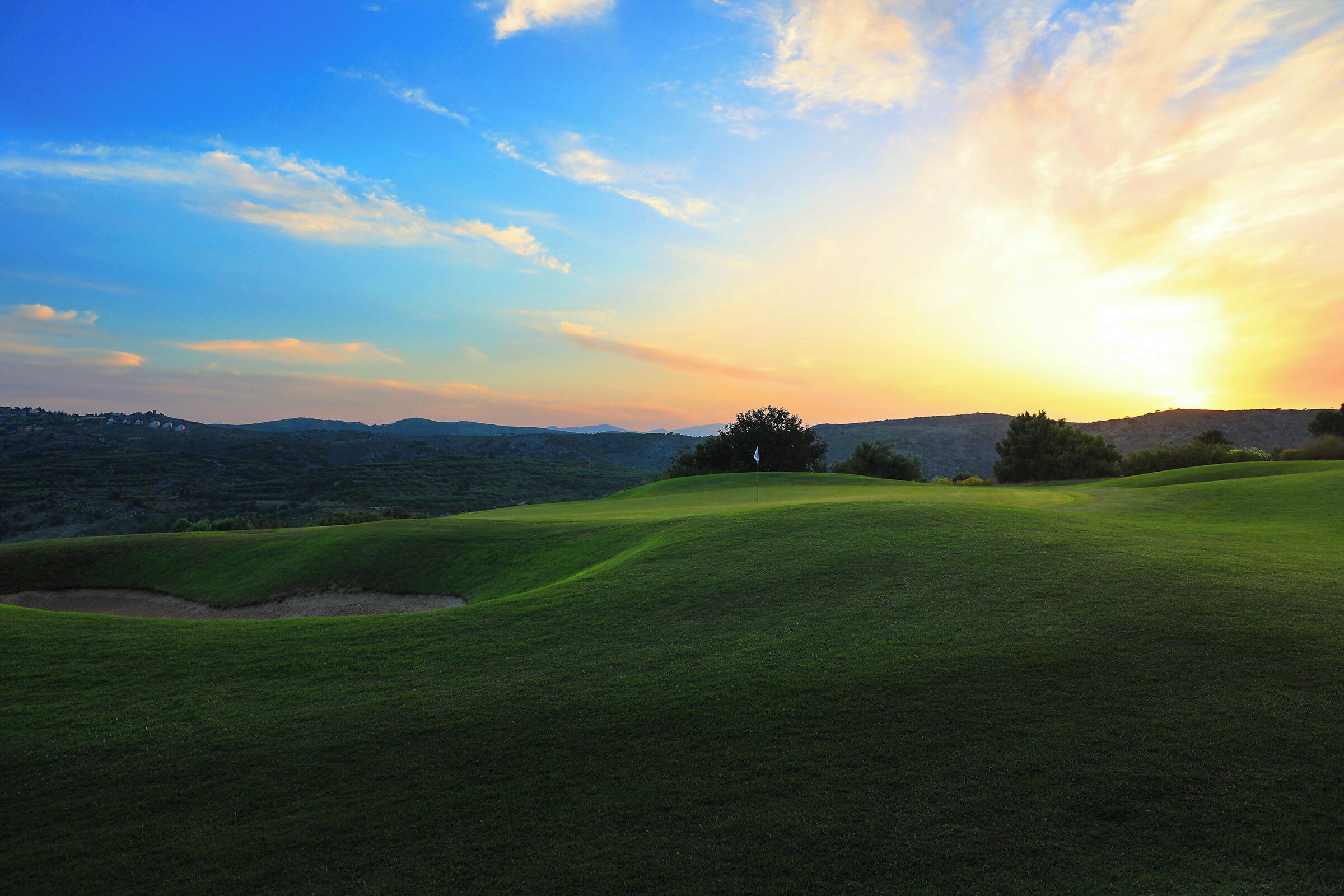 A golf course with a flagstick, rolling hills, and a sunset in the background.