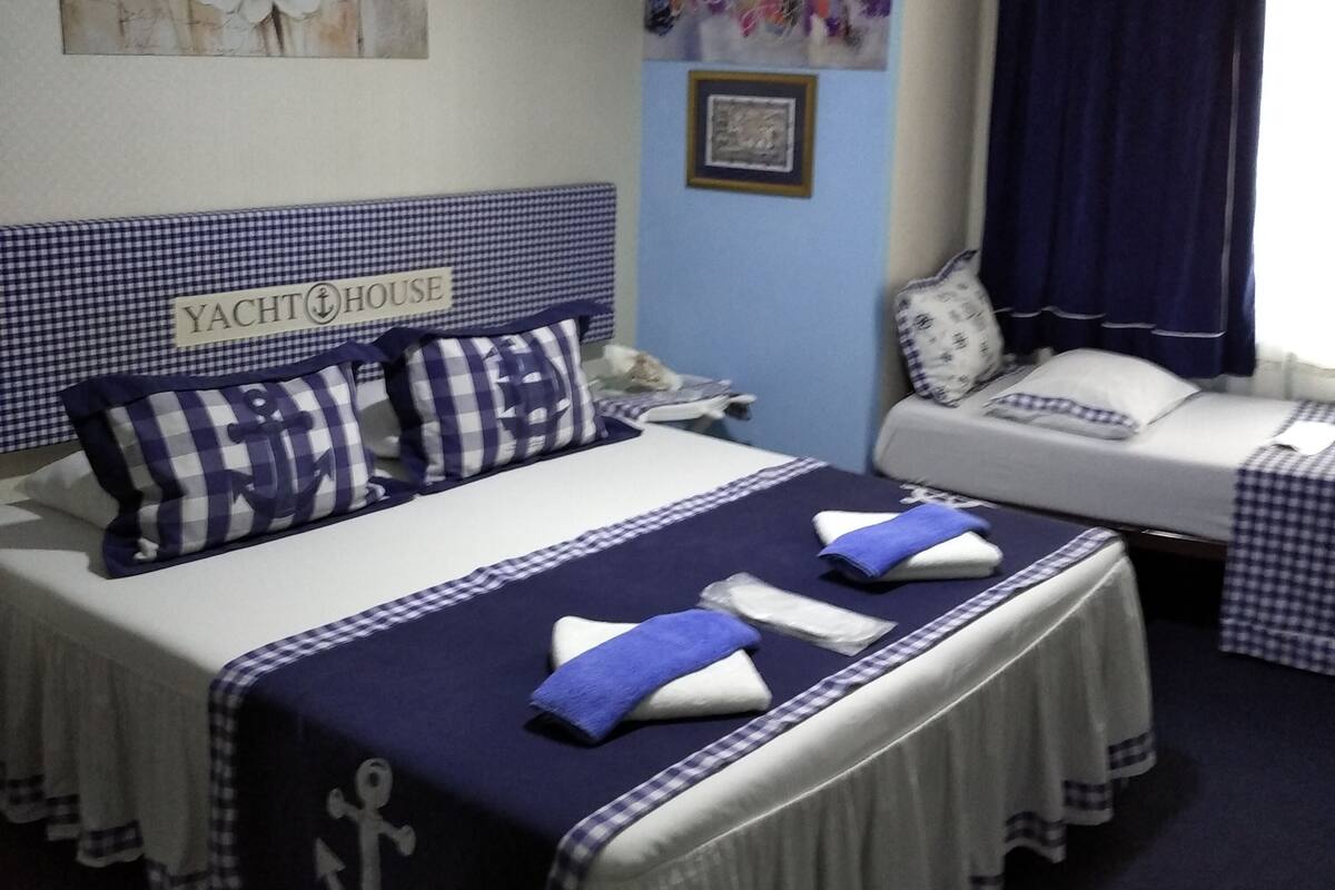 A hotel room with a bed featuring a checkered bedspread and pillows, a bedside table with a lamp, and a wall with a framed picture and a sign that reads "YACHT HOUSE".
