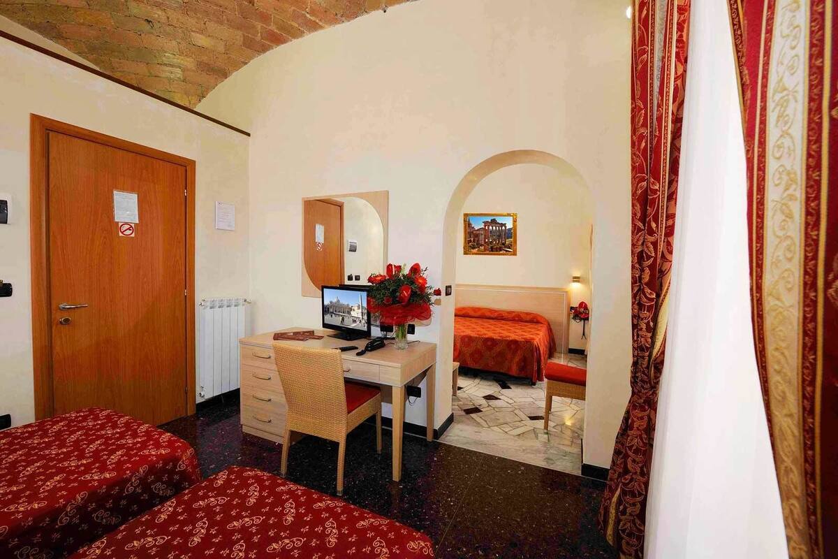 A hotel room with a wooden desk, a computer, a bed with red bedding, a wicker chair, and a small table with a vase of flowers.