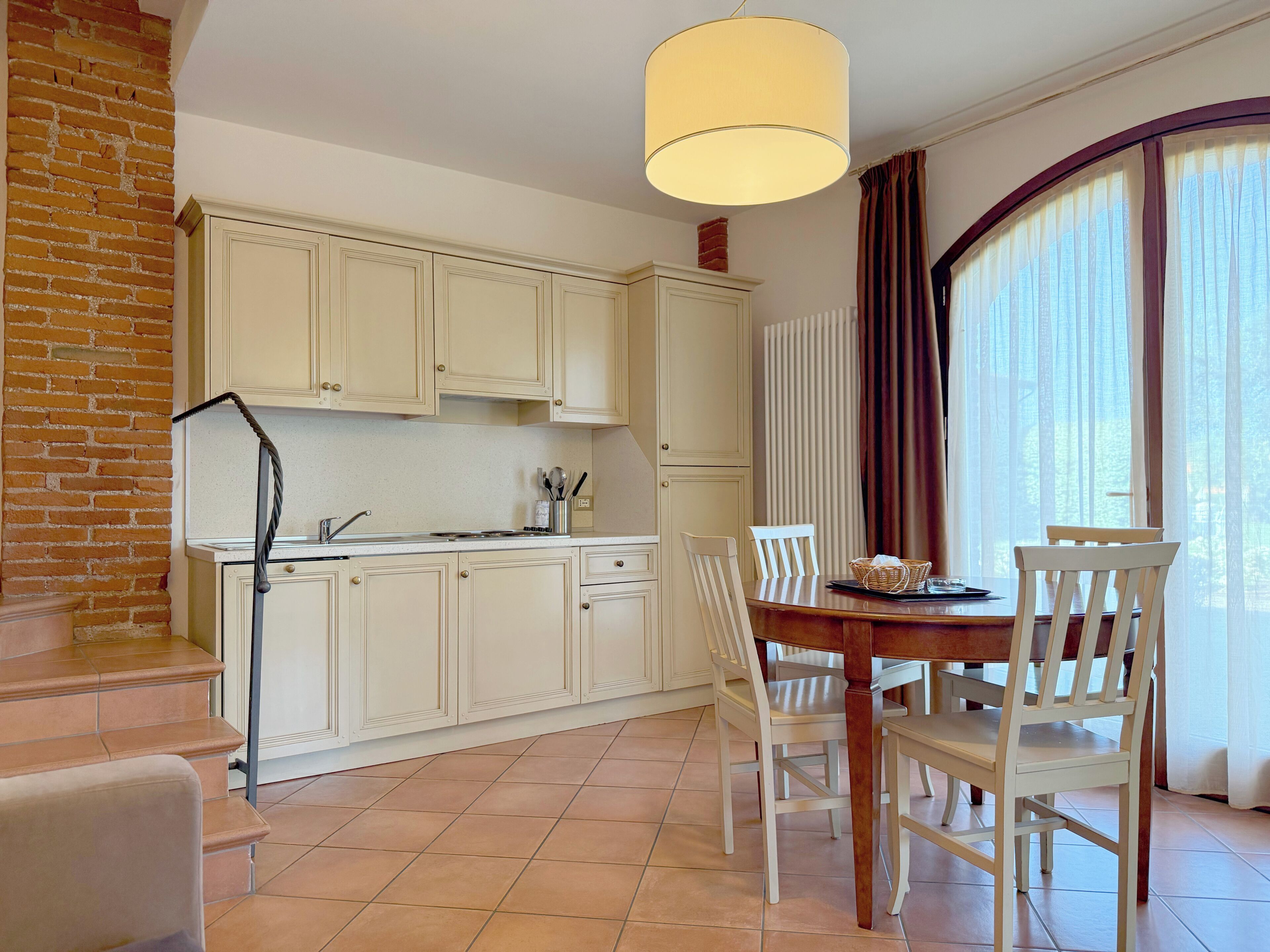 Una cocina con armarios blancos, una mesa de comedor con sillas, una lámpara colgante y una ventana con cortinas.