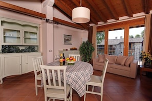 A dining area with a table set for two, a white cabinet with glass doors, a beige sofa, and a large window with curtains. - Residence Borgo Mondragon (Lazise)