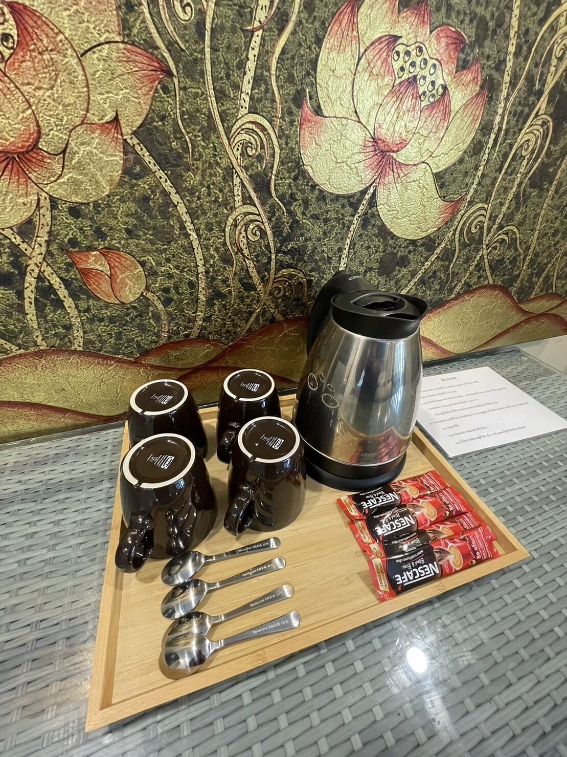 A tray with four black coffee mugs, a stainless steel coffee pot, a set of spoons, and packets of Nescafe instant coffee on a wooden tray.