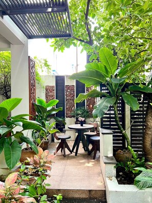 Ein überdachter Patio-Bereich mit rundem Tisch und Stühlen, umgeben von verschiedenen Kübelpflanzen und einer Zierwand.