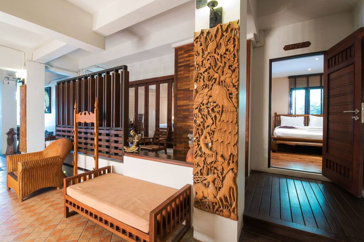 A room with a bed, a wooden chair, a decorative wall panel, and a view of another room.