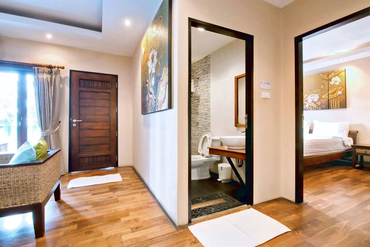 A hotel room with a wooden floor, a bed, a bathroom with a toilet and sink, and a door leading to another room.