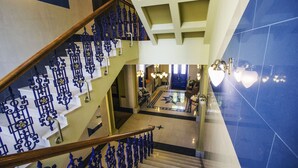 Una imponente escalera con barandillas de metal decoradas lleva a un vestíbulo con piso estampado y una lámpara de cristal.