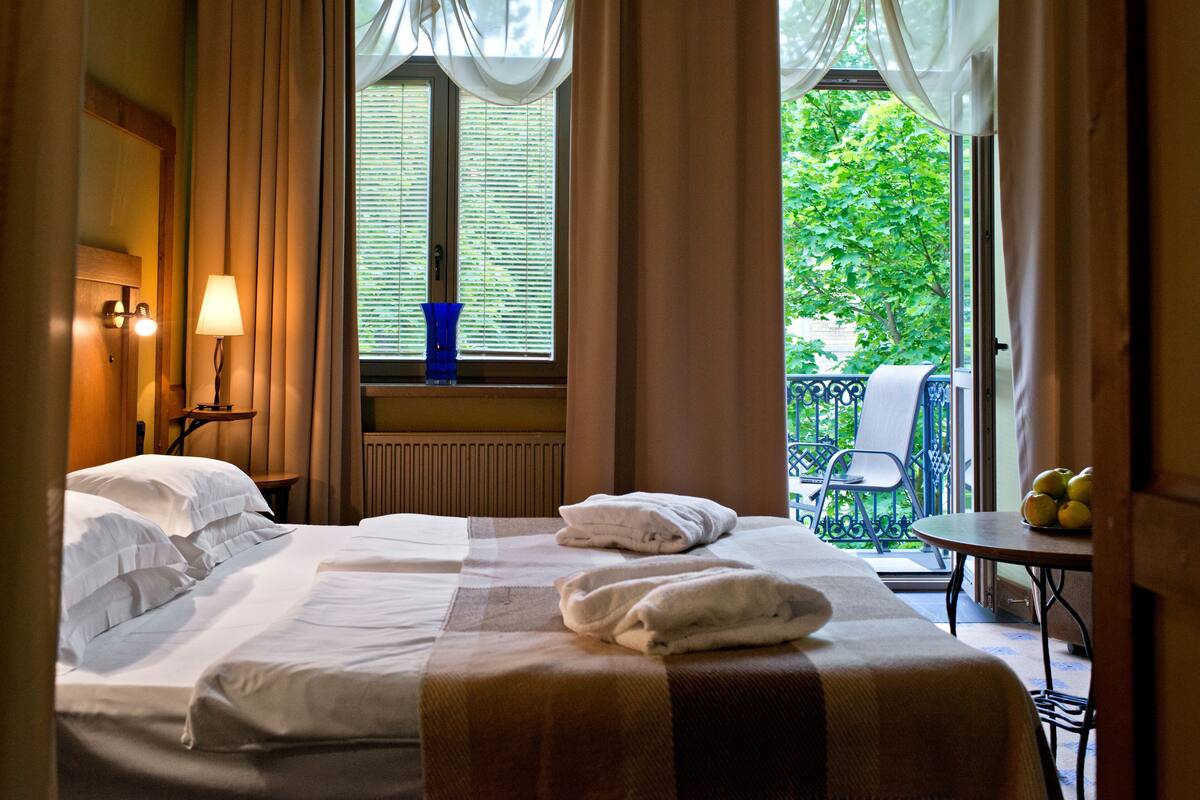 Et hotelværelse med to senge, et vindue med persienner, et bord med frugt og en balkon med en stol.