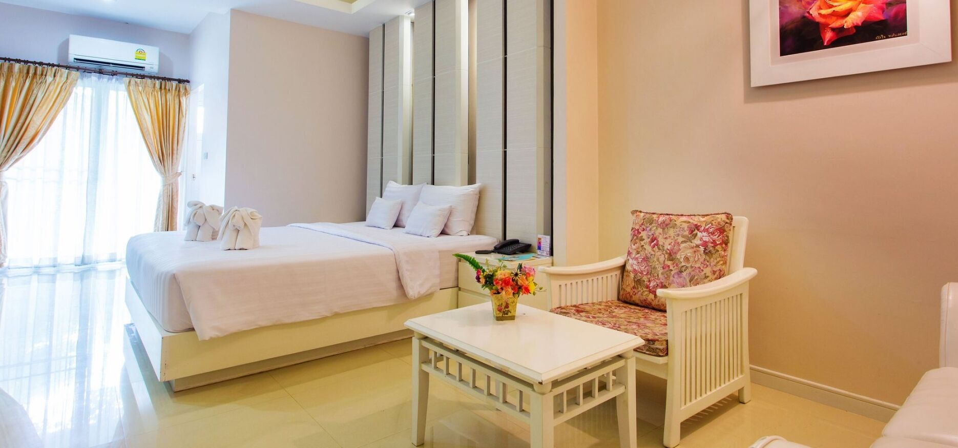 Une chambre d’hôtel avec un lit, une chaise, une petite table sur laquelle est posé un vase de fleurs, et un tableau accroché au mur.