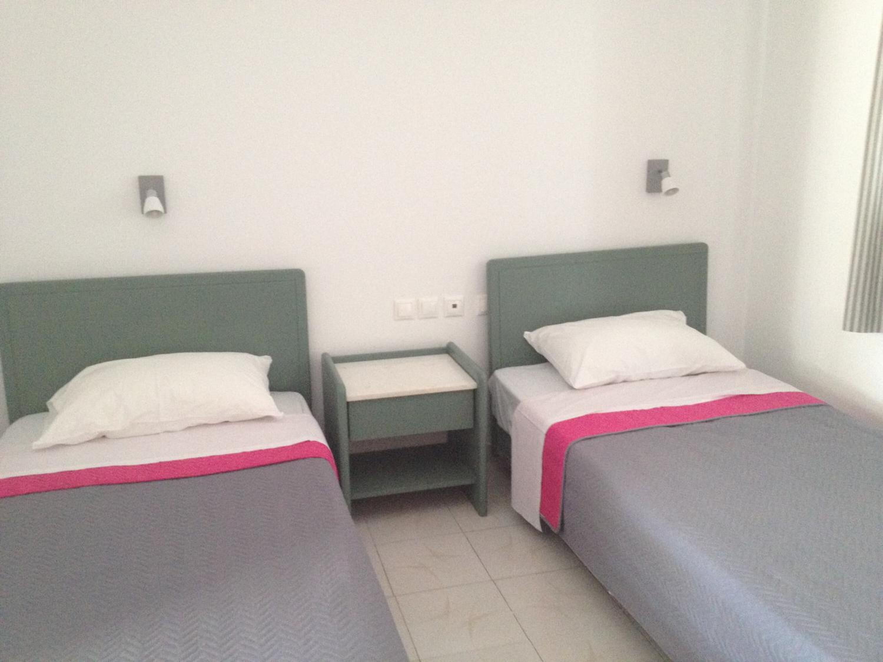 Una habitación de hotel con dos camas individuales, cada una con cabecero verde y almohadas blancas. Hay una mesita de noche pequeña con una lámpara entre las camas.