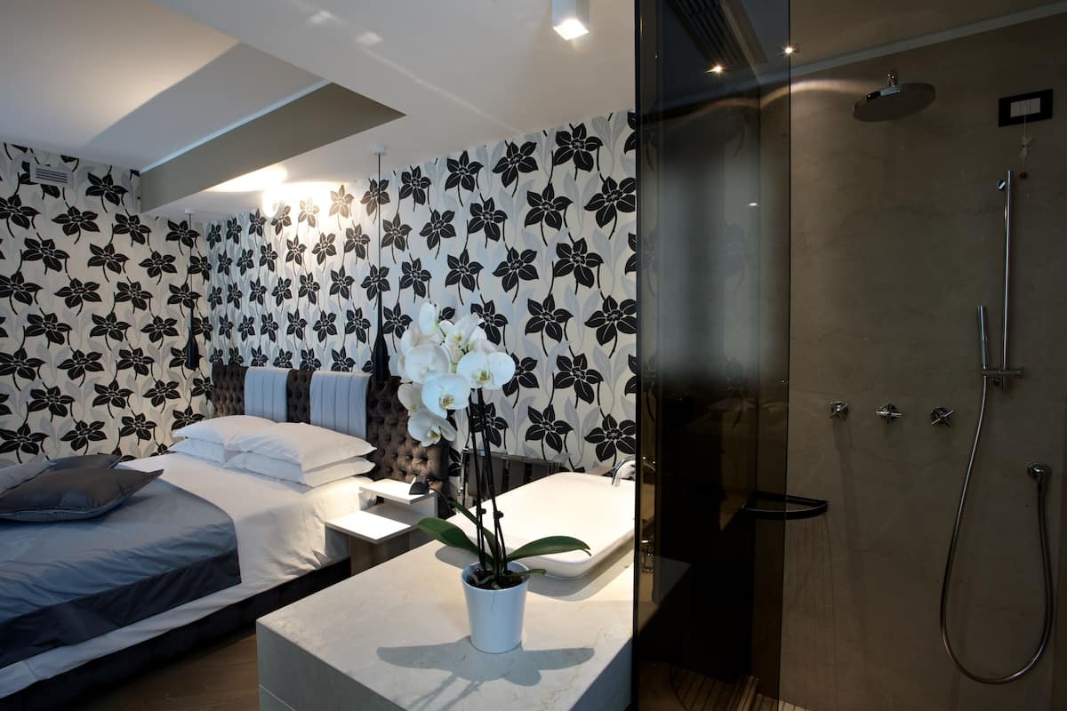 A modern hotel room with a bed, a small table with a potted orchid, a shower area, and a bathroom with a white sink and mirror.