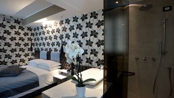 Quarto de hotel moderno com cama, uma pequena mesa com uma orquídea em vaso, área de chuveiro e banheiro com pia e espelho brancos.