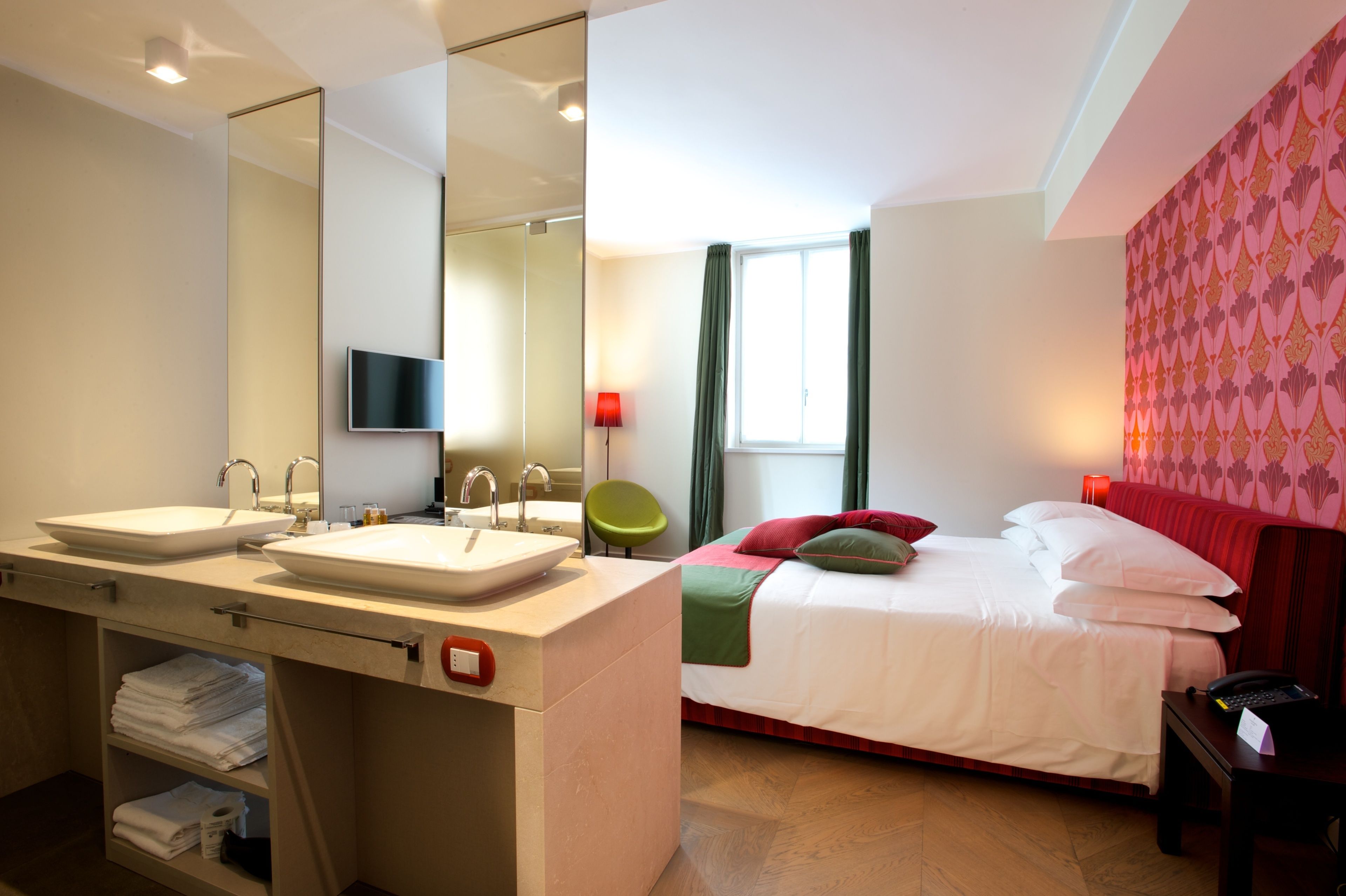 A modern hotel room with a large mirror, two sinks, a bed with red and green bedding, and a television mounted on the wall.
