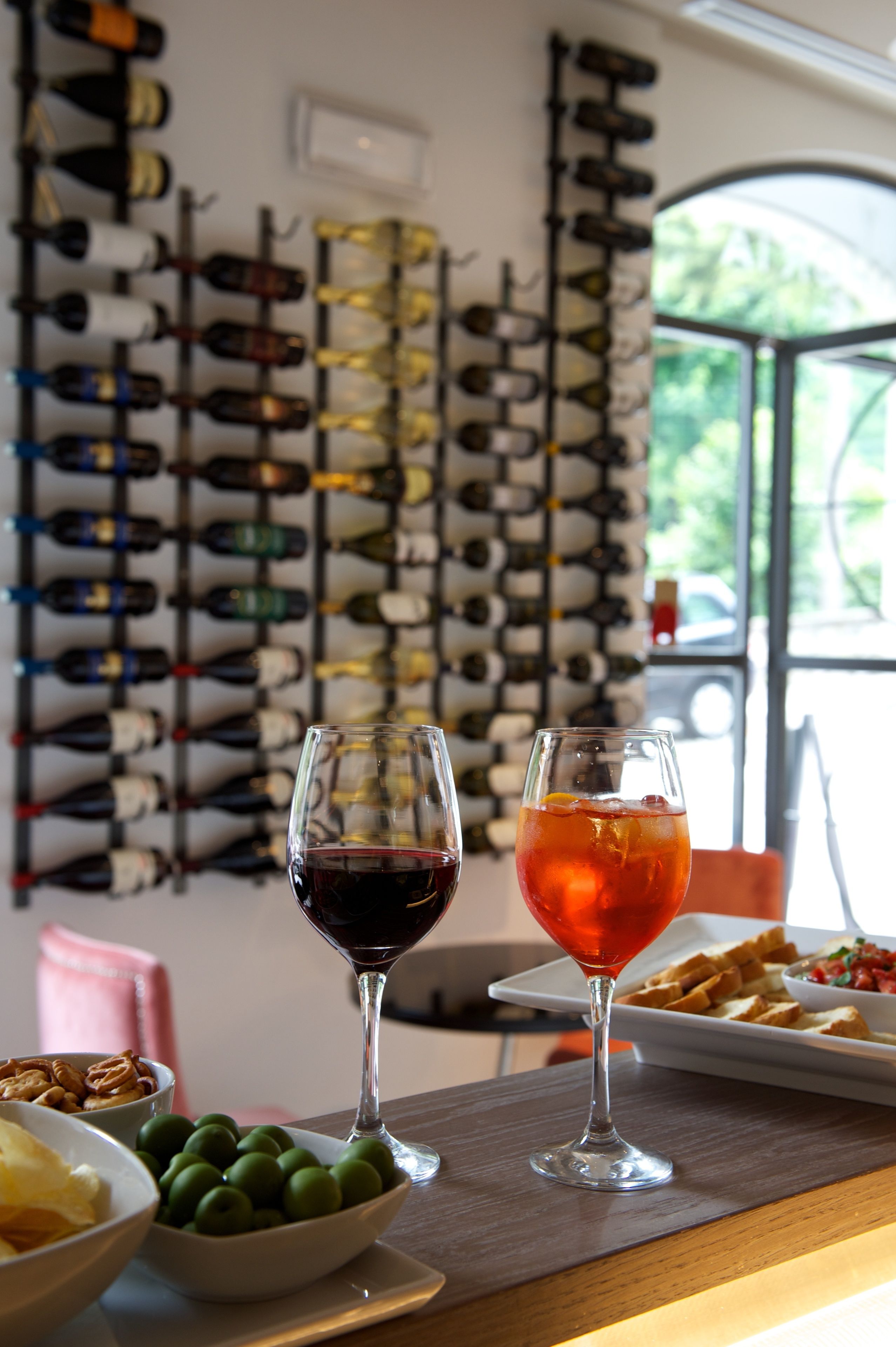 A wine rack with various bottles, two glasses of wine, a bowl of olives, and a plate of bread on a table.