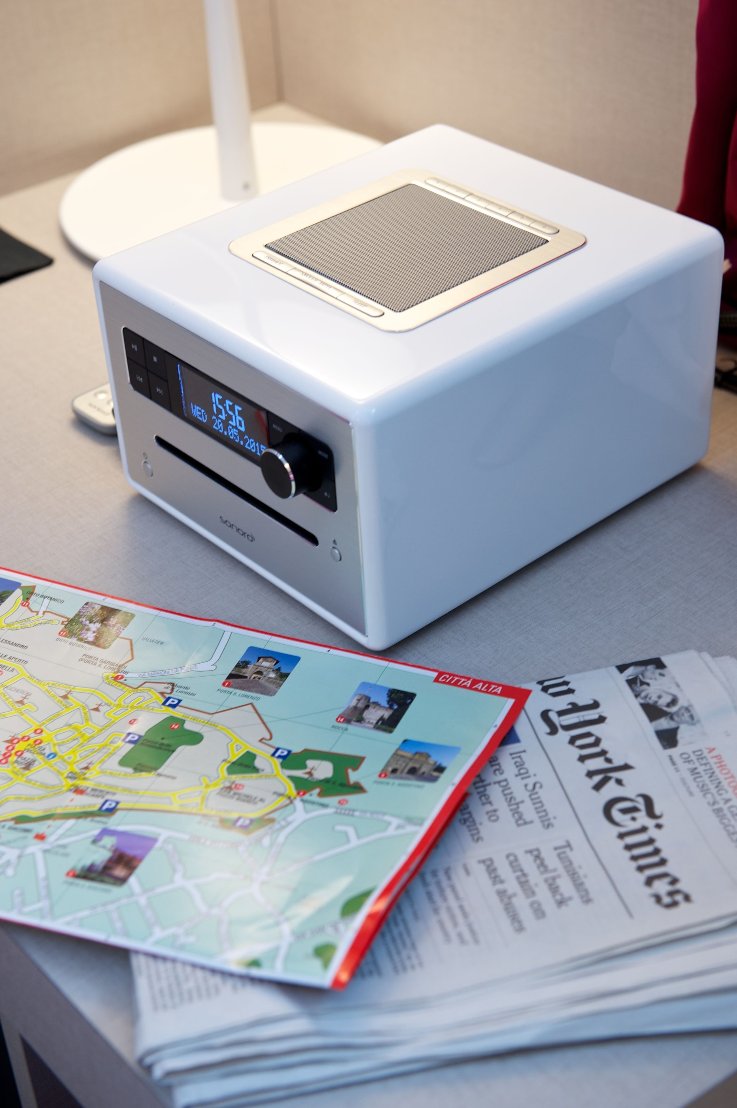 A white portable speaker with a digital display, a map, and a newspaper on a table.
