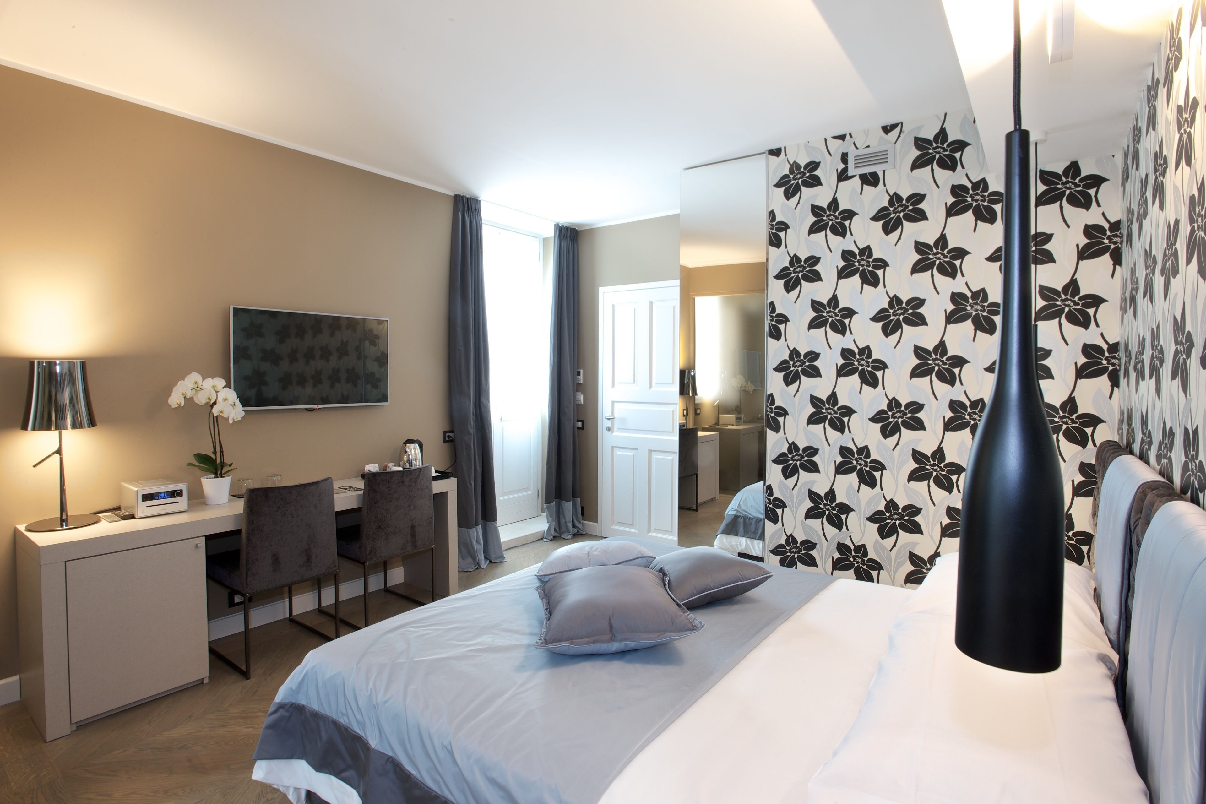 Una habitación de hotel moderna con cama, escritorio y sillas. Hay un espejo grande, una planta en maceta y un florero en el escritorio.