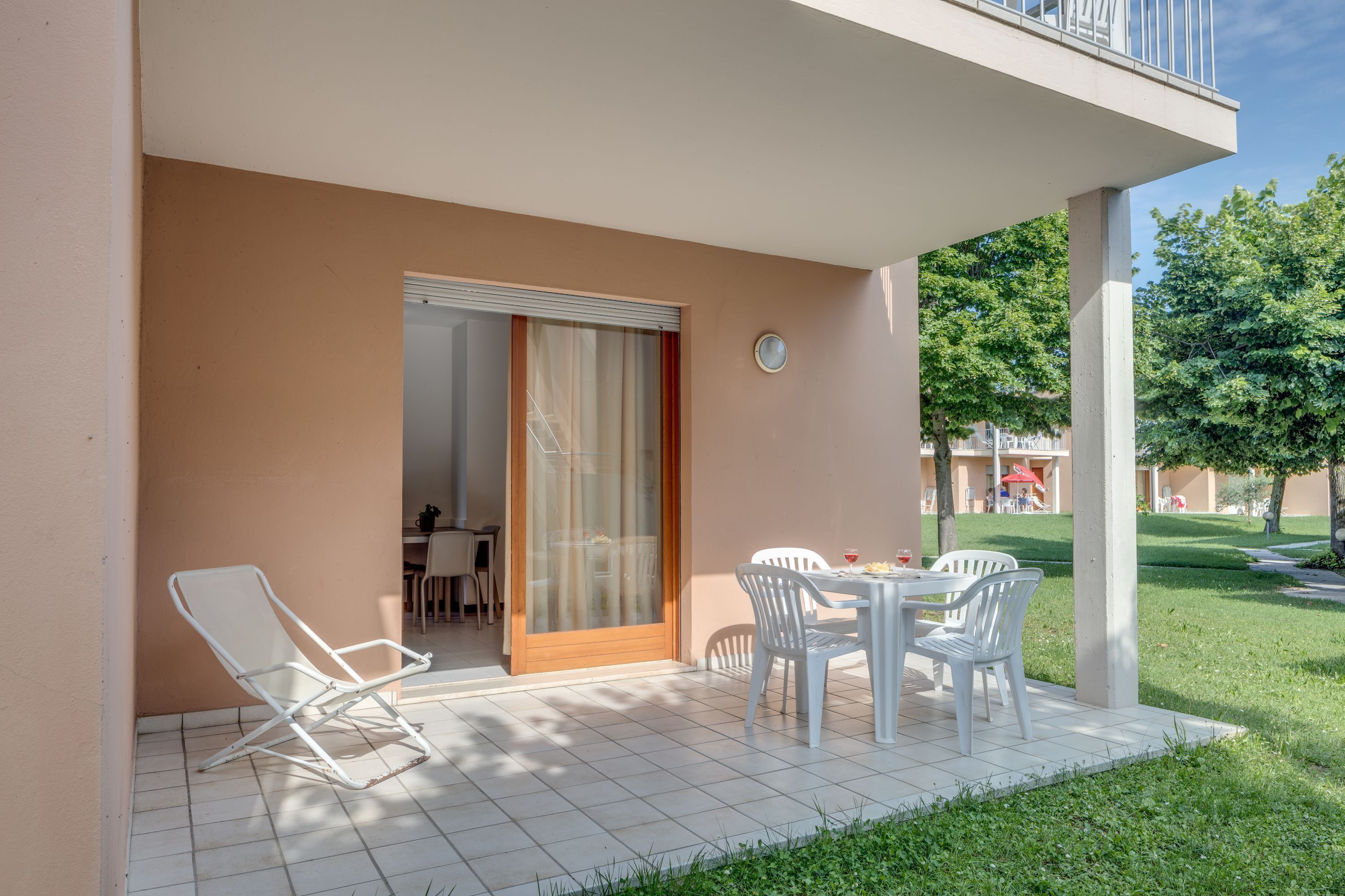 Un espacio exterior cubierto con una mesa y sillas, una silla lounge blanca y un ventanal corredero que da a un espacio interior.
