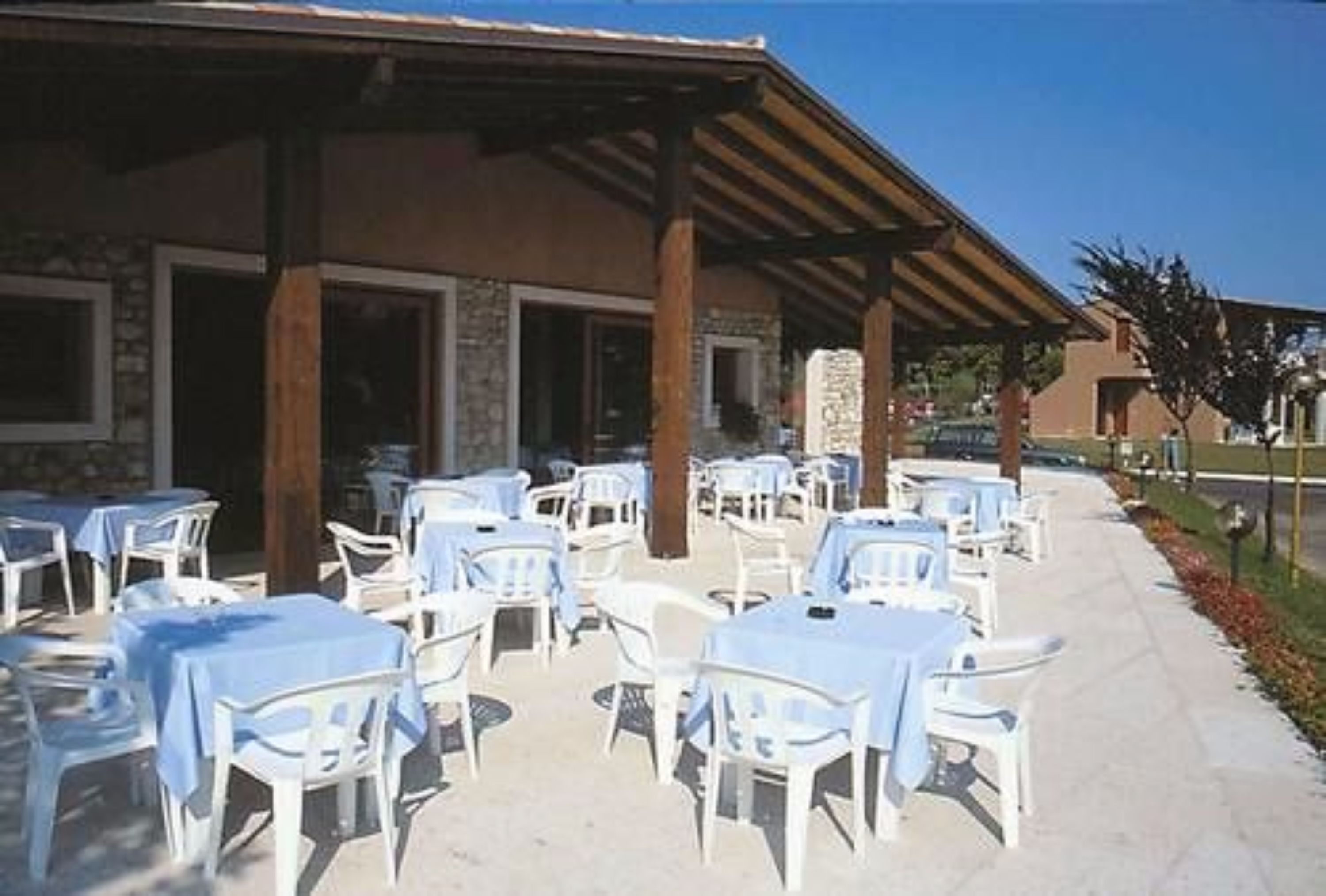 An outdoor patio area with white plastic tables and chairs, covered by a wooden pergola.