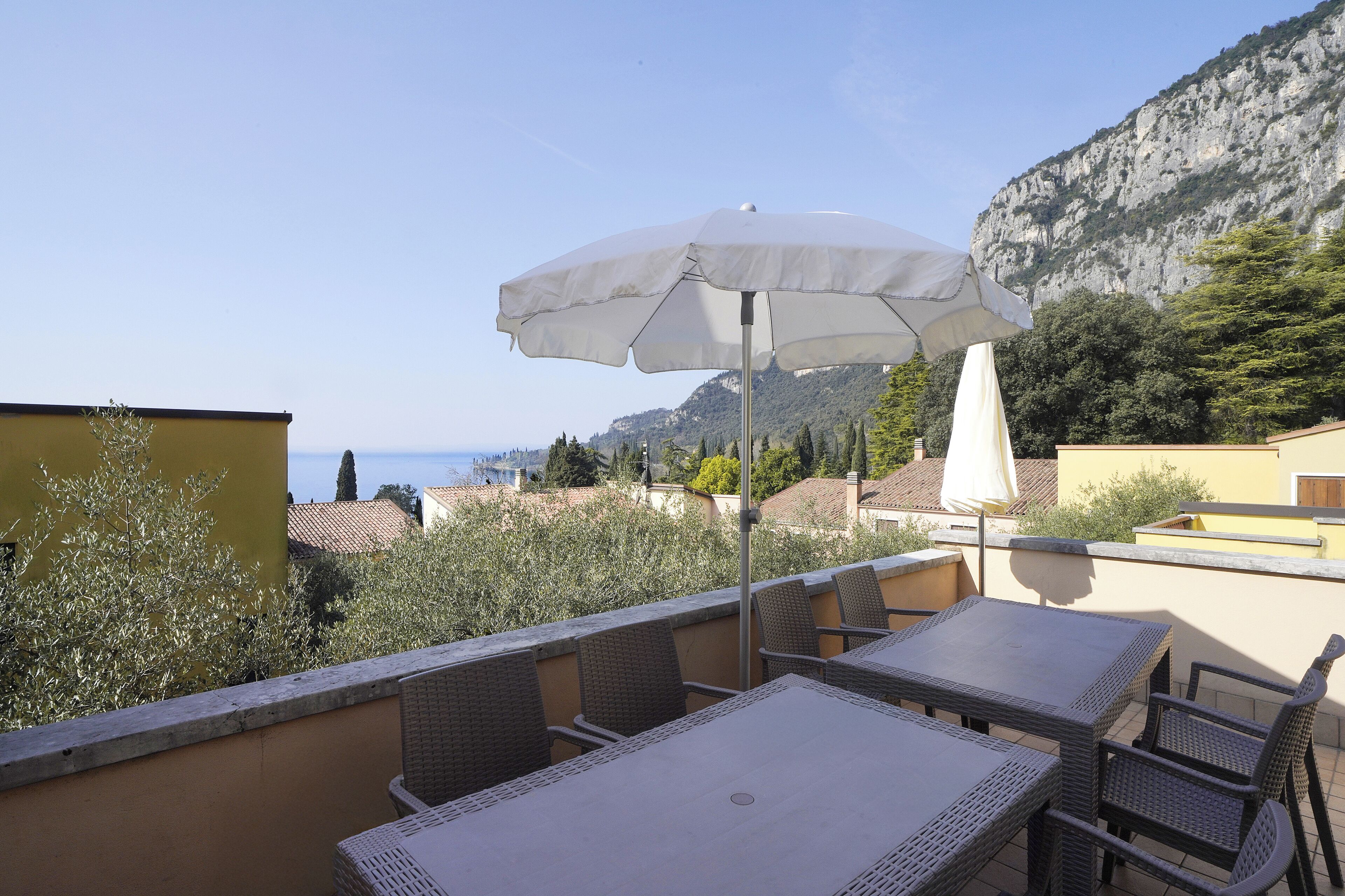 A terrace with a table and chairs, a white umbrella, and a view of the sea and mountains.