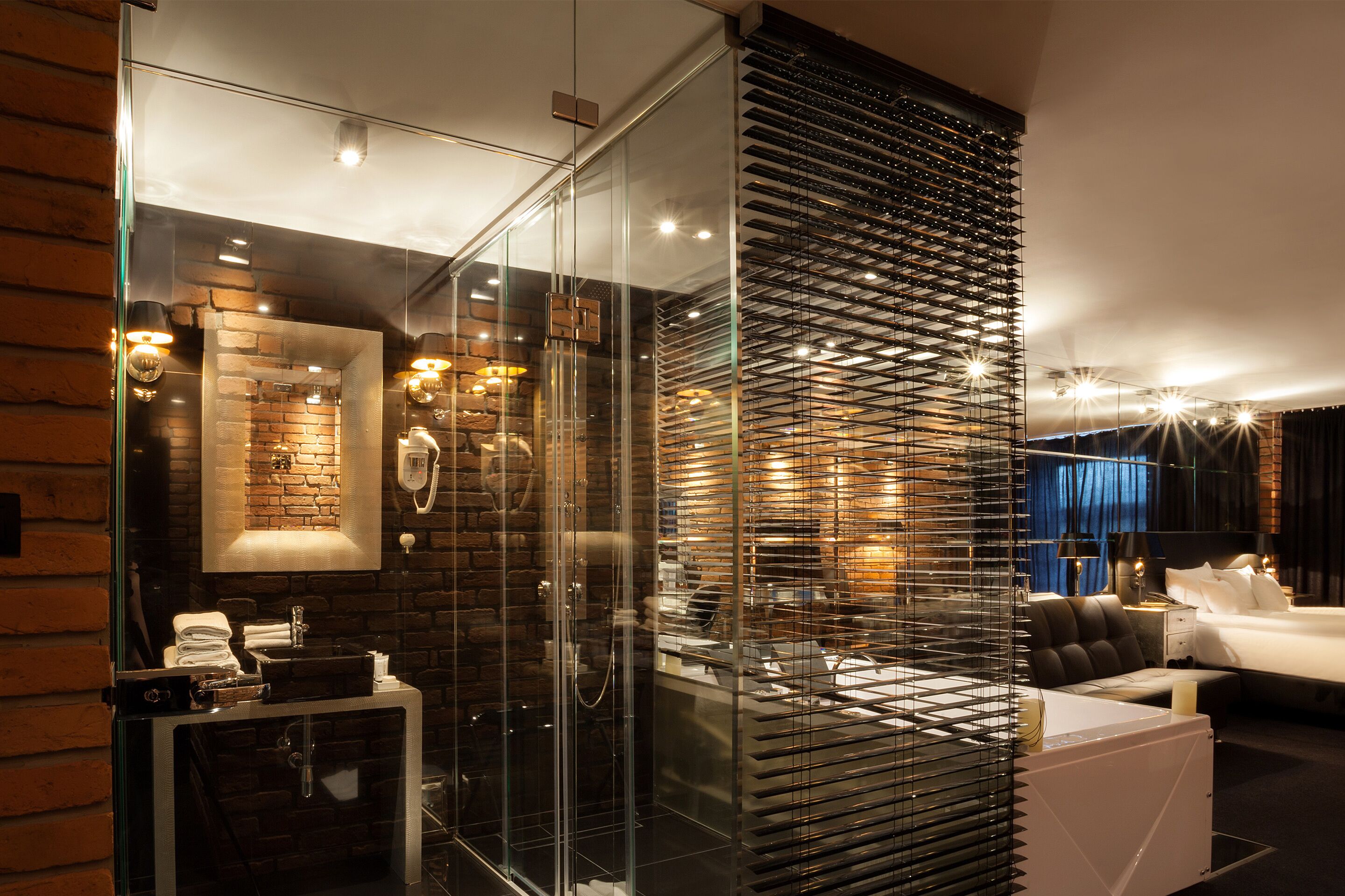 A modern hotel room with a glass partition, a brick wall, a bed, a sofa, and a small table with a lamp.