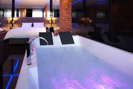 A modern hotel room with a large bathtub, a brick accent wall, and a large mirror reflecting the room.