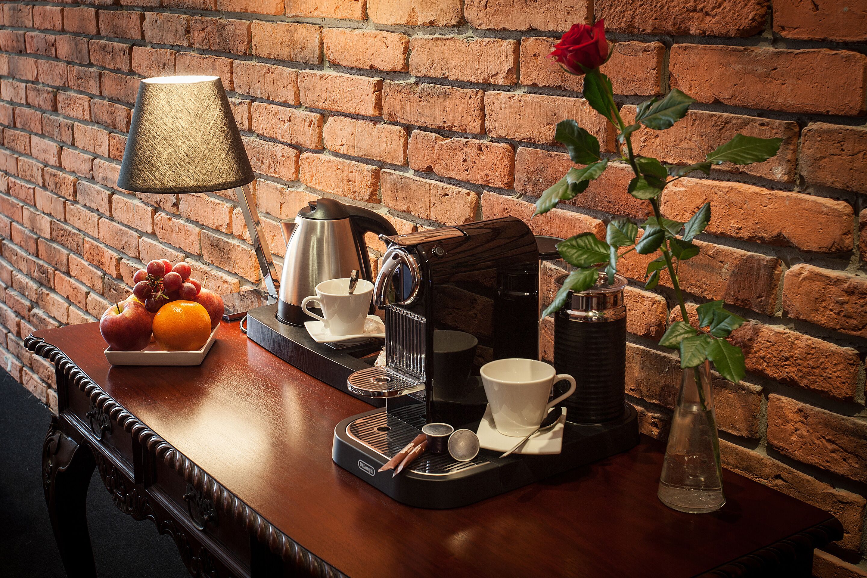 Una mesa de madera con una cafetera, tazas, un jarrón con una rosa y un frutero.