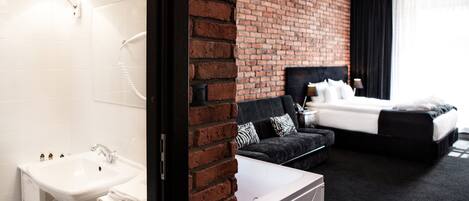 A hotel room with a white sink, a black sofa, a bed with black bedding, and a brick wall.