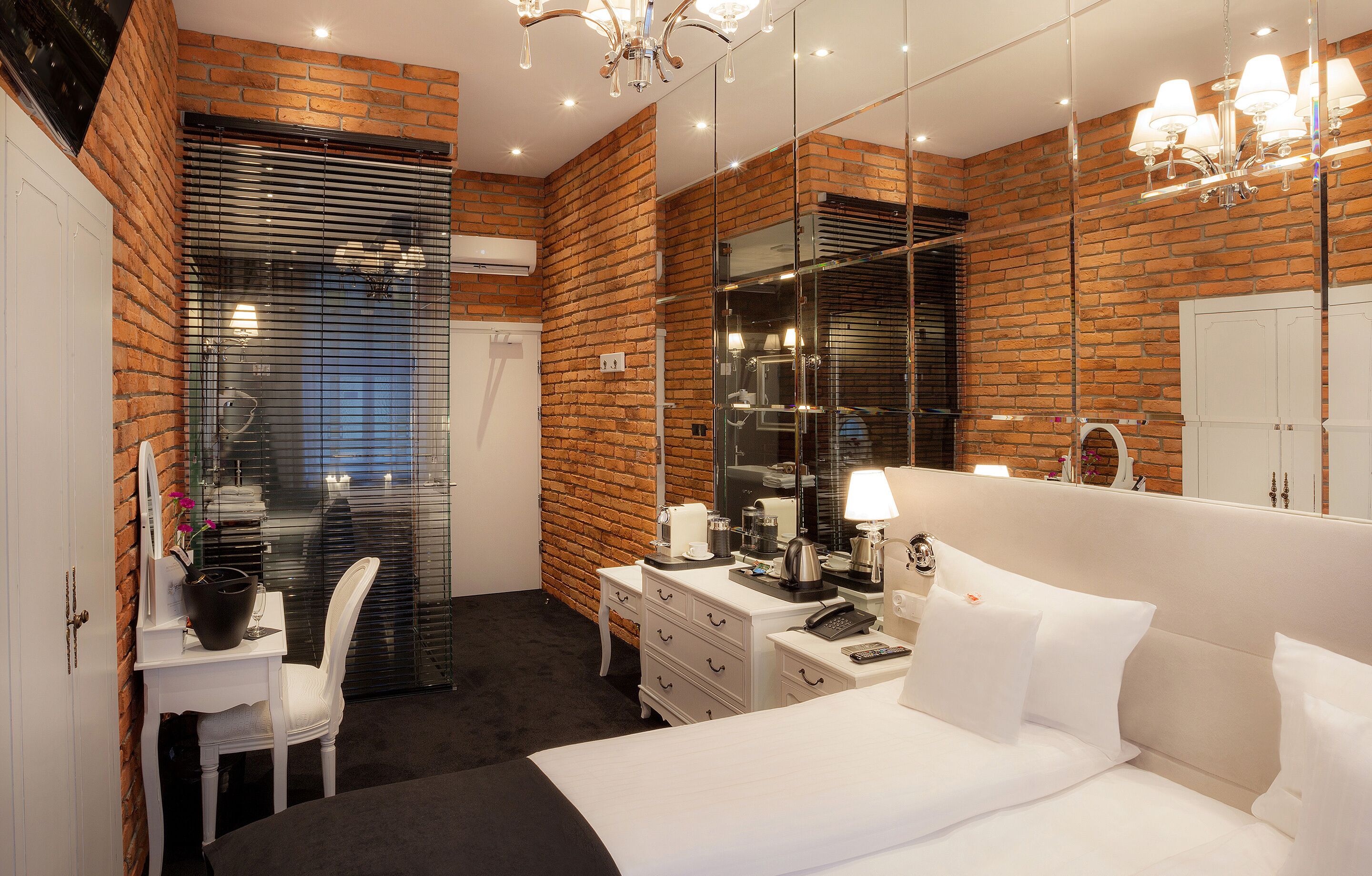 Een moderne hotelkamer met een bakstenen muur, een grote spiegel, een wit bed, een bureau met een stoel en een kaptafel met een spiegel.