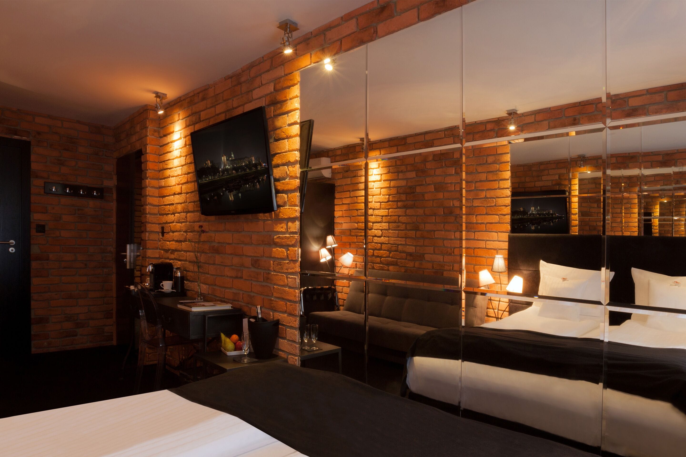 A hotel room with a brick wall, a bed, a sofa, a TV, and a small table with a coffee machine.