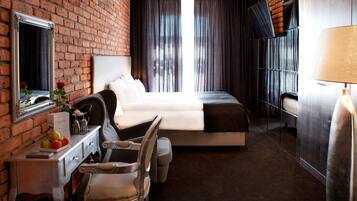A hotel room with a brick wall, a bed, a desk with a mirror, a chair, a lamp, and a window with curtains.