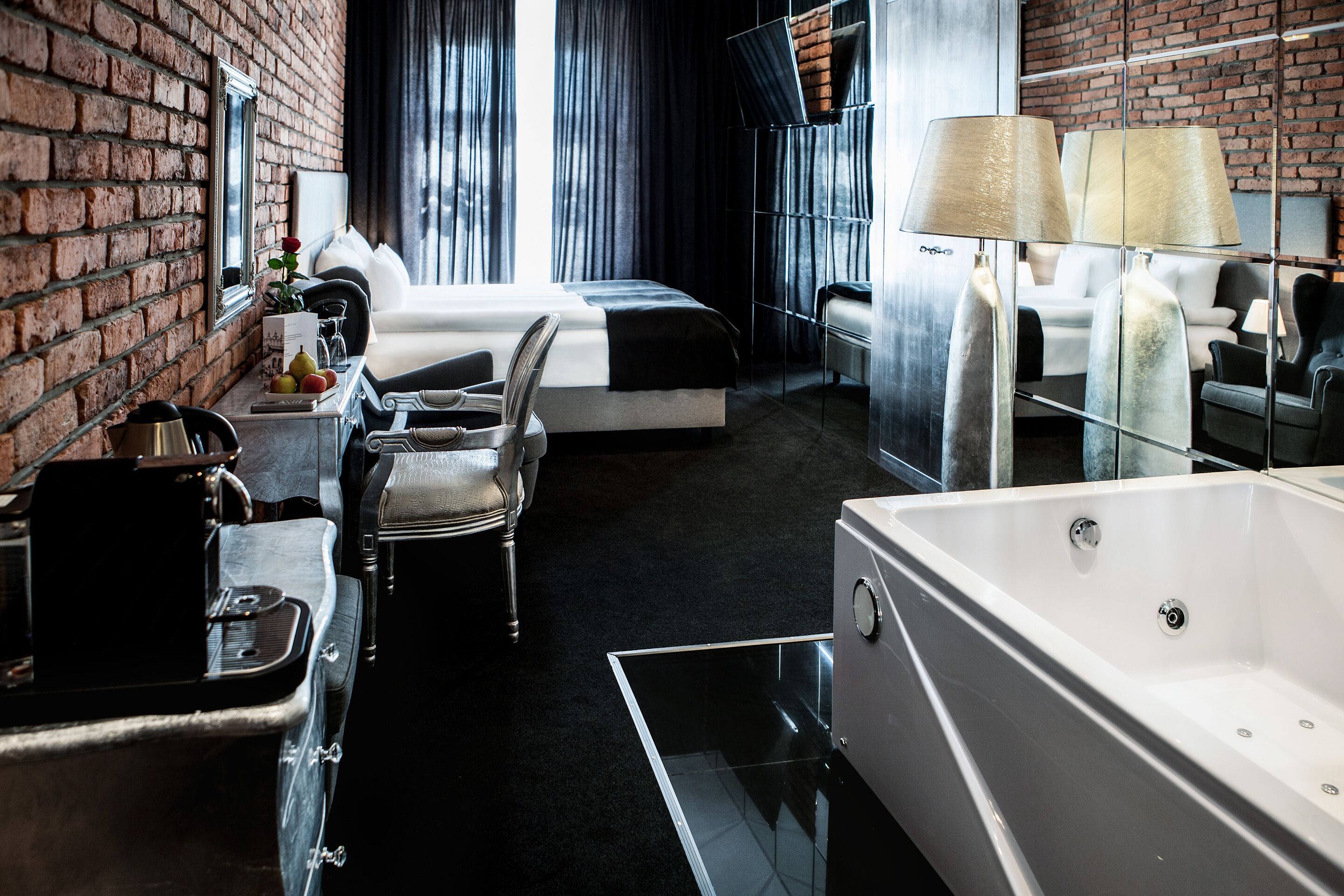 A modern hotel room with a brick wall, a large window with curtains, a bed, a desk with a chair, a bathtub, and a lamp.