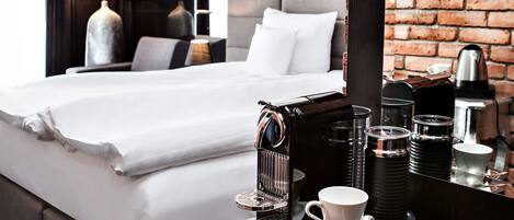 Habitación de hotel con una cama bien hecha, una cafetera, tazas y un espejo que refleja una pared de ladrillo.