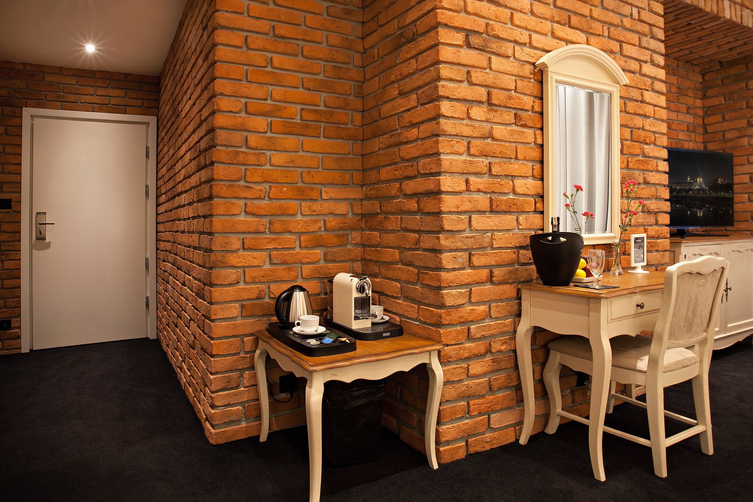 Um quarto com parede de tijolos, porta branca, uma mesa pequena com uma cafeteira, uma cadeira e uma televisão na parede.