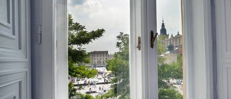 A view through a window with white trim and a wooden handle, overlooking a public square with people and buildings, including a prominent tower.