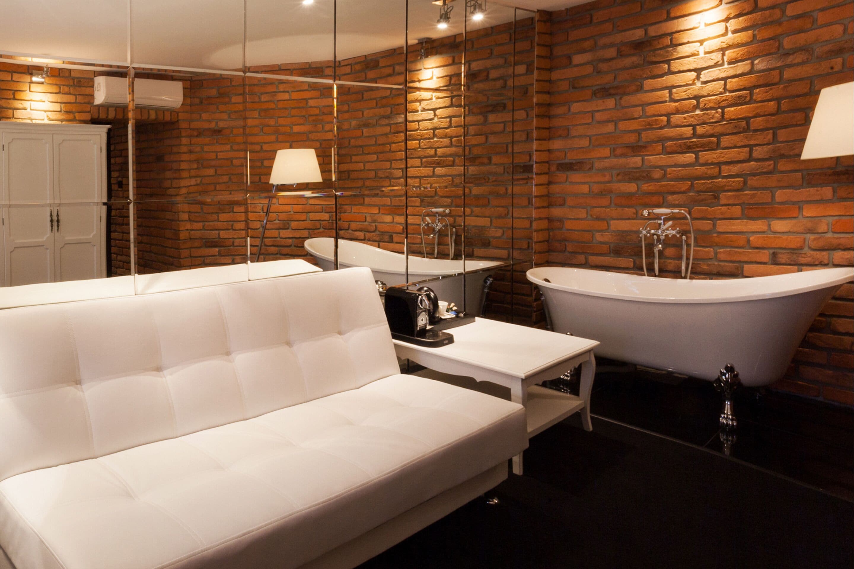 A modern bathroom with a white bathtub, a white sofa, and a mirror reflecting the brick wall.