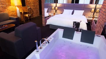 Ett modernt hotellrum med en stor säng, en sittgrupp med soffa och fåtölj, ett litet bord med ett fat med frukt och ett stort badkar.