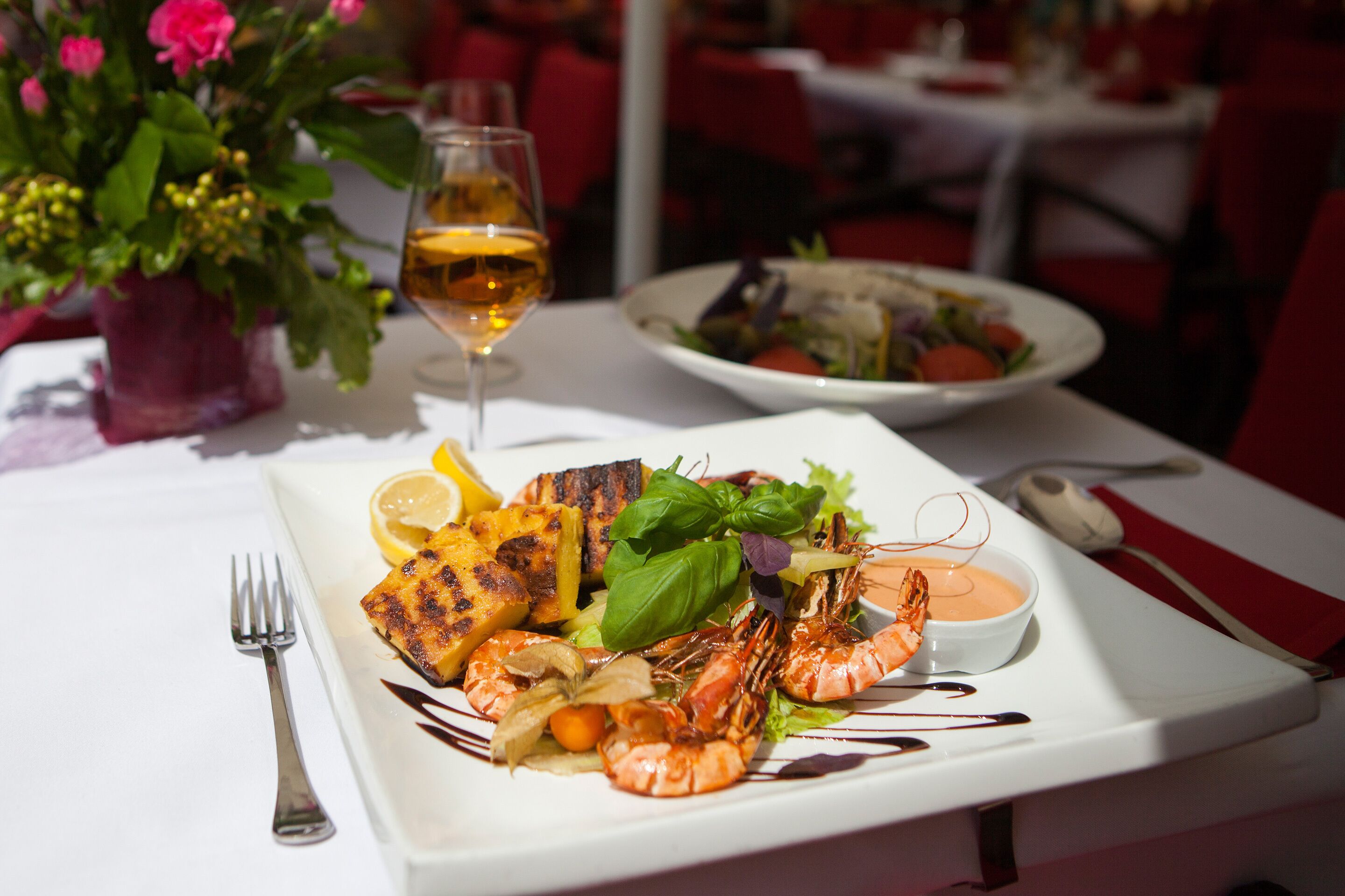 A dining table with a plate of grilled food, a glass of wine, and a bowl of sauce.