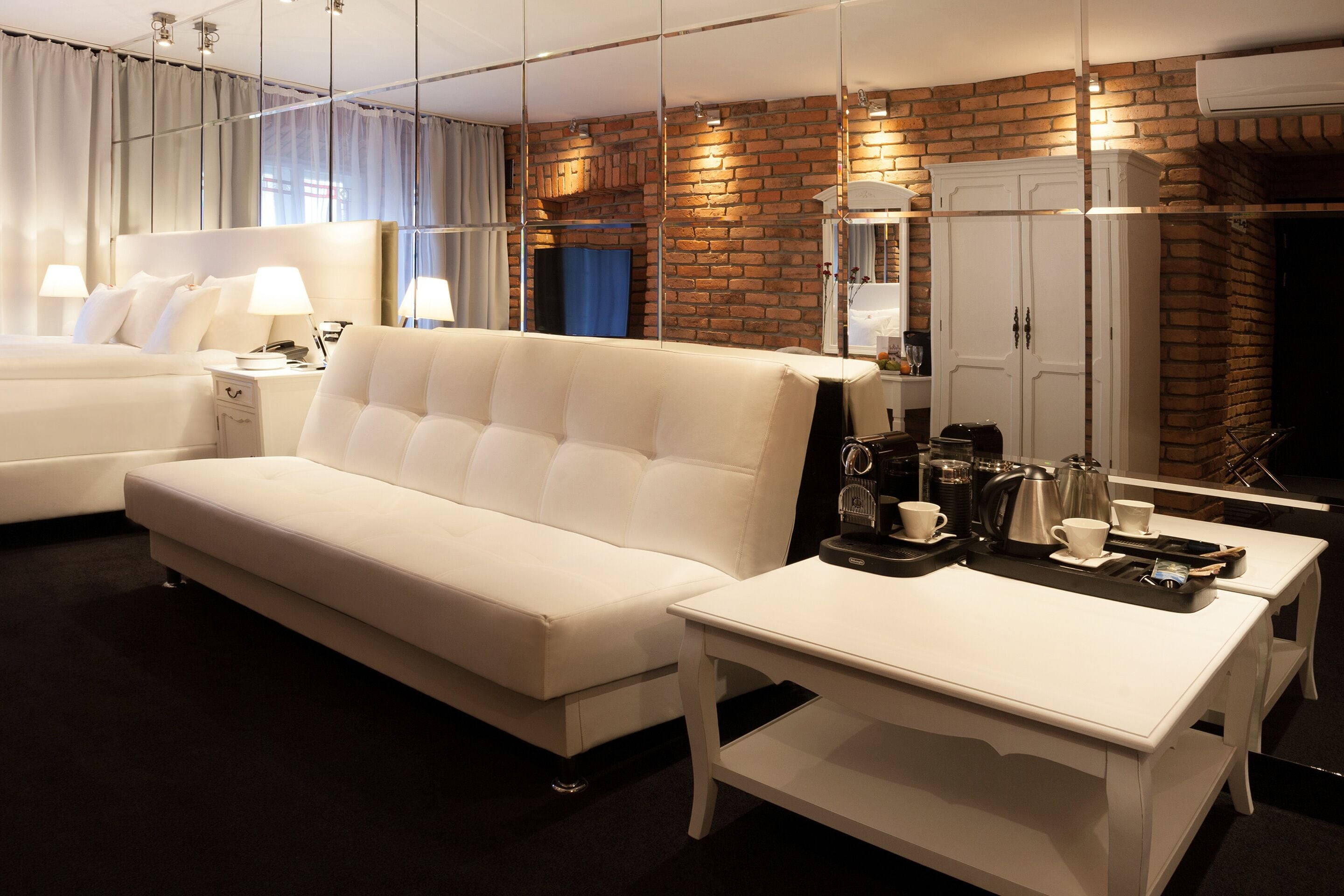 Habitación de hotel con un sofá blanco, una pequeña mesa blanca con una bandeja de alimentos para el desayuno, una cama con ropa de cama blanca y una pared de ladrillo con un espejo y un armario.