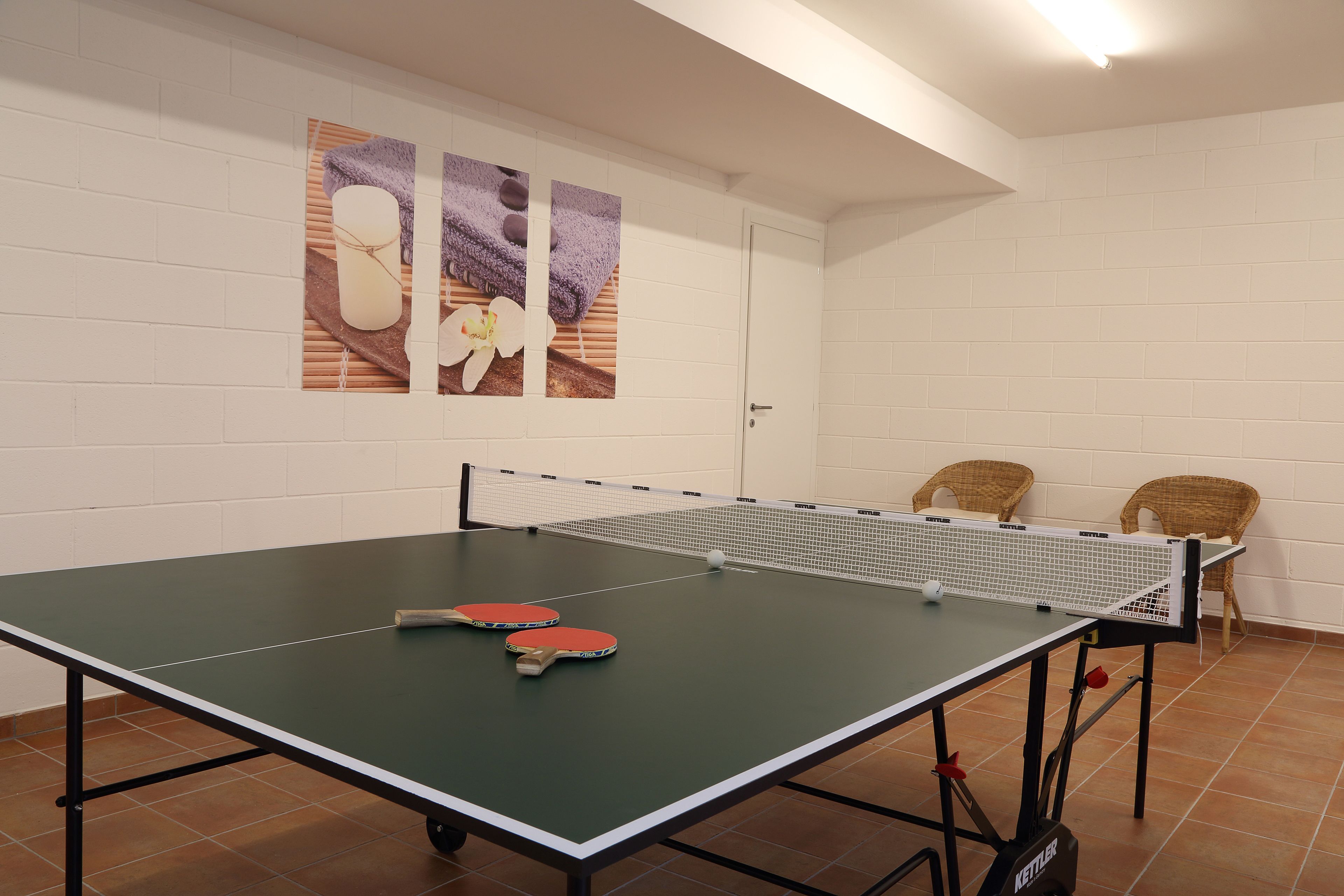 A ping pong table with two paddles and a ball, two wicker chairs, and two wall art pieces depicting flowers and towels.