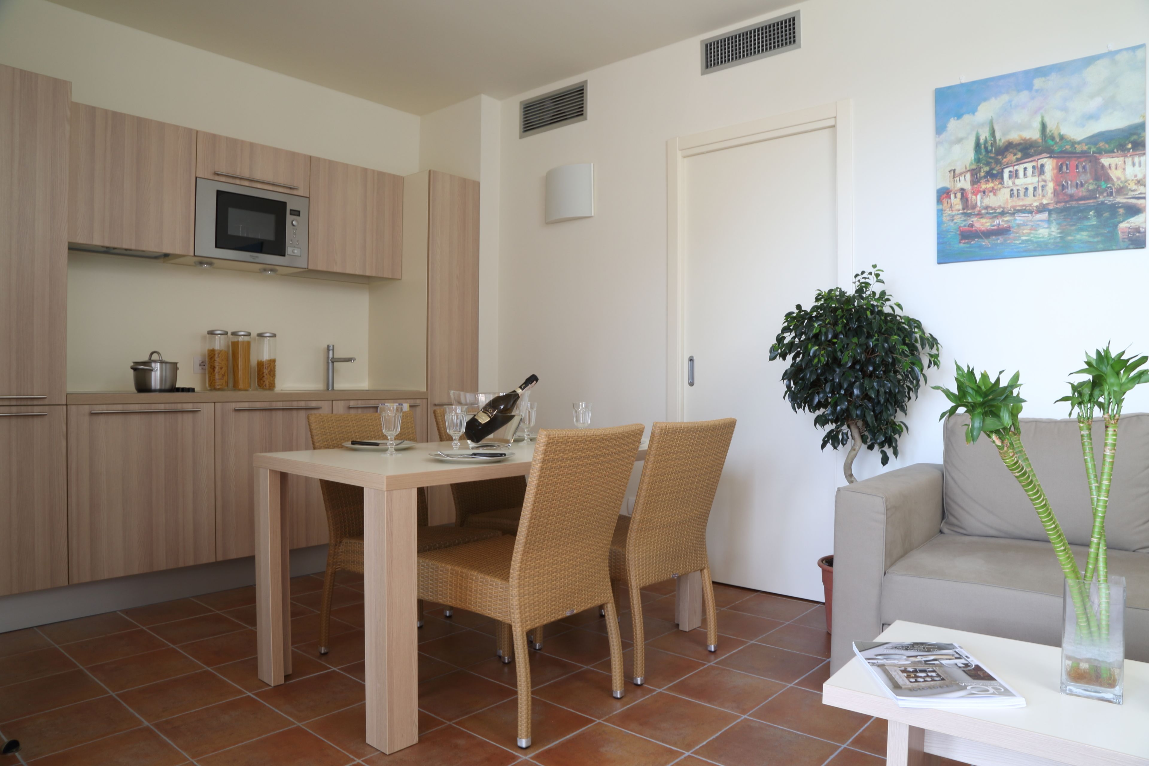 A modern kitchen with wooden cabinets, a microwave, and a dining area with wicker chairs. There is a potted plant and a painting on the wall.