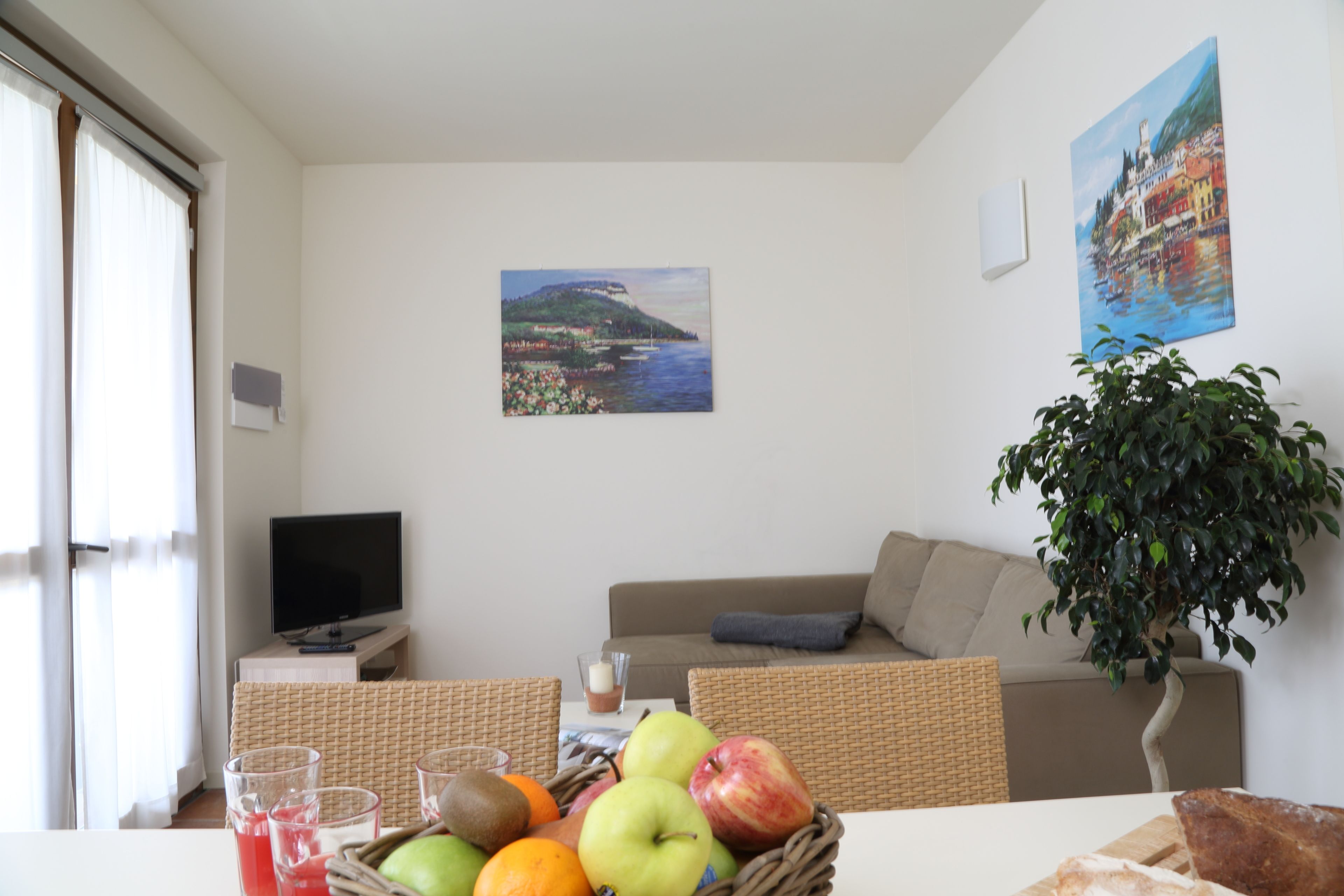 Uma sala de estar com televisão, um sofá, uma mesa de jantar com frutas e copos, e duas pinturas na parede.