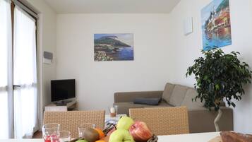 Uma sala de estar com televisão, um sofá, uma mesa de jantar com frutas e copos, e duas pinturas na parede.