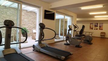 Ein Fitnessraum mit Laufbändern, Crosstrainern und einem Ergometer. An der Wand ist ein Fernseher angebracht und das Fenster bietet einen Blick nach draußen.