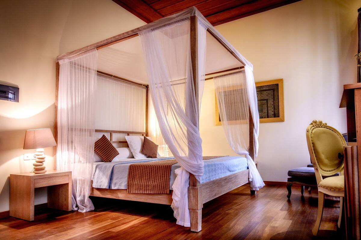 Una cama con dosel, una mesita de noche con lámpara y una silla en una habitación con suelo de madera y un cuadro en la pared.
