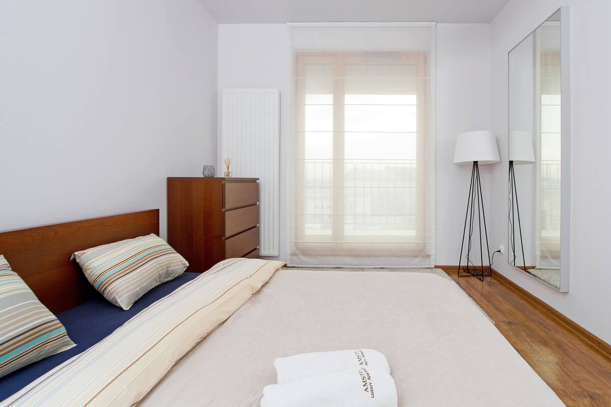 Ein Schlafzimmer mit Bett, Kommode, Spiegel und einem Fenster mit durchsichtigen Vorhängen.