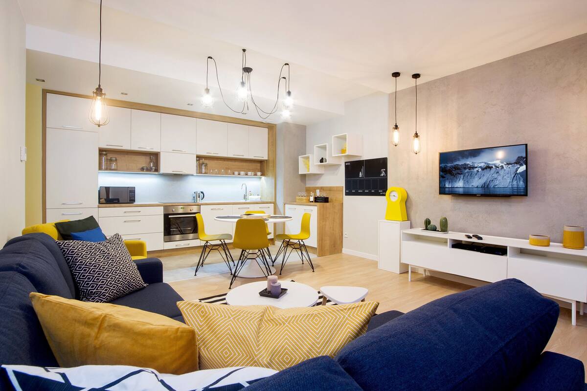 Ein modernes Wohnzimmer mit einer Couch, einem Esstisch und Stühlen. Es gibt eine Küchenzeile mit Schränken und einem an der Wand befestigten Fernseher.